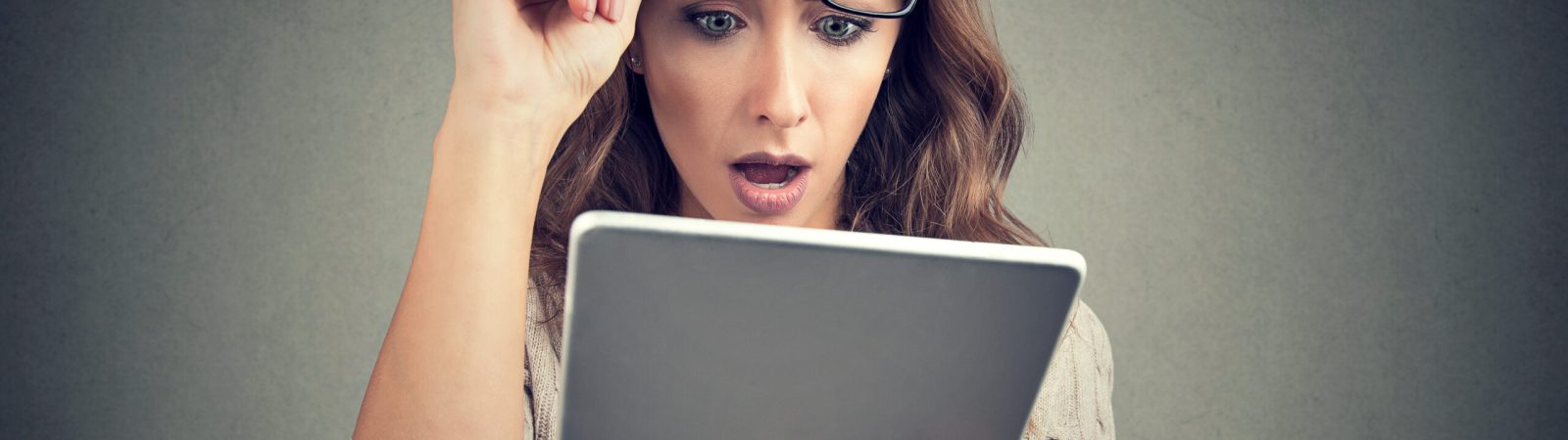 Young girl in eyeglasses watching tablet with shock having bad news and posing on gray.