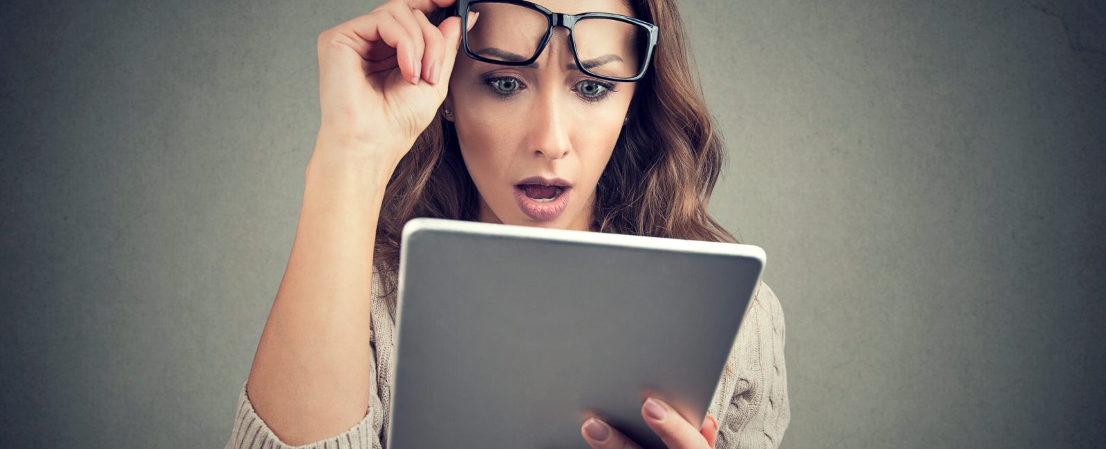 Young girl in eyeglasses watching tablet with shock having bad news and posing on gray.