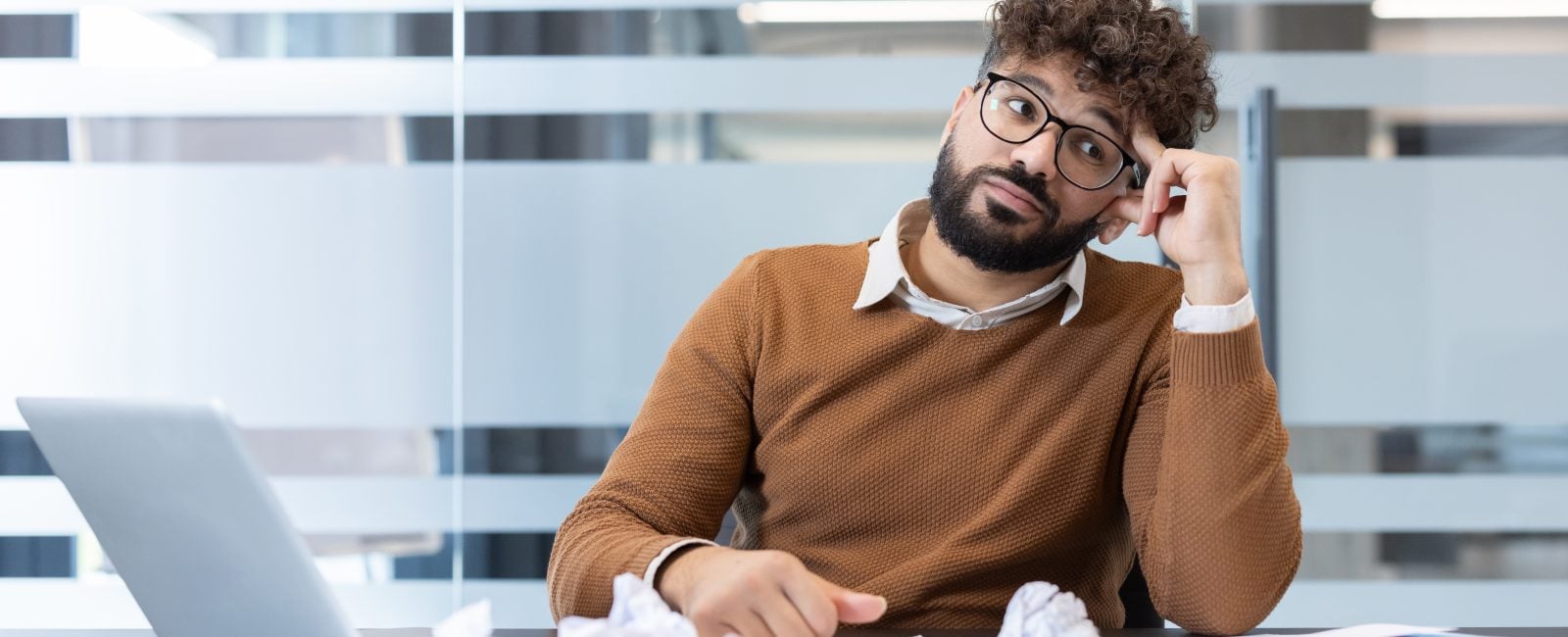 Man experiencing mental block at office desk, surrounded by crumpled paper, laptop, and documents, highlighting stress, challenge, and creative struggle in the workspace