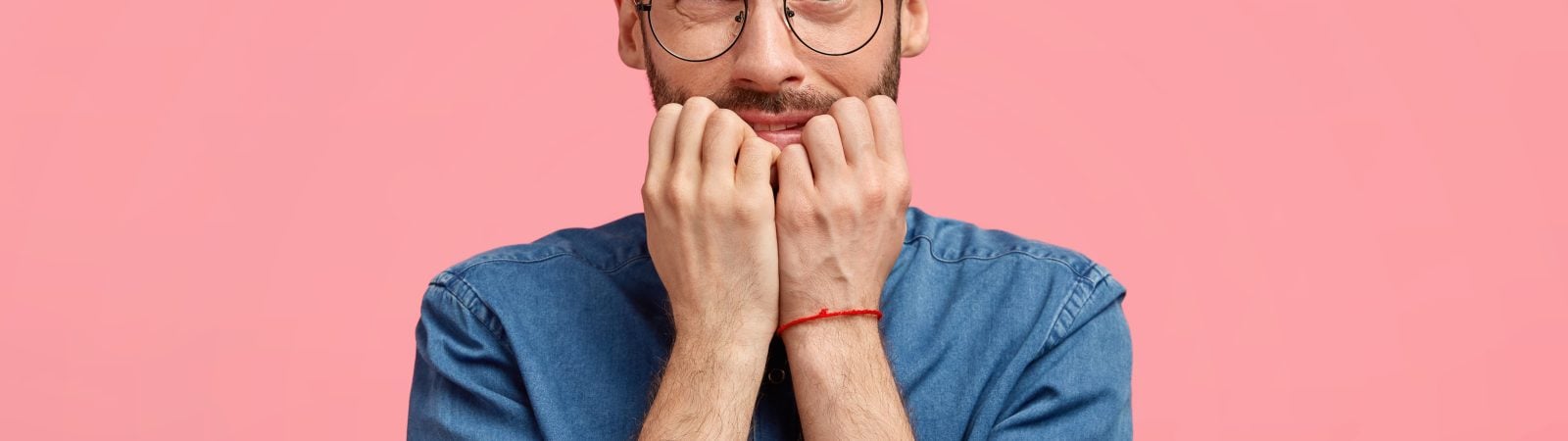 Attractive young man with confused nervous expression, bites finger nails, worries to make terrible mistake, feels anxious, looks embarrassed at camera, wears denim shirt, isolated over pink wall