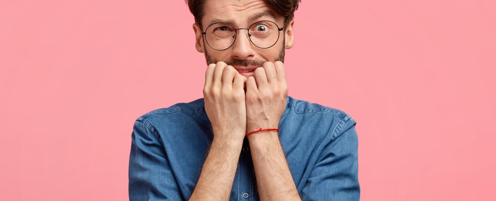 Attractive young man with confused nervous expression, bites finger nails, worries to make terrible mistake, feels anxious, looks embarrassed at camera, wears denim shirt, isolated over pink wall