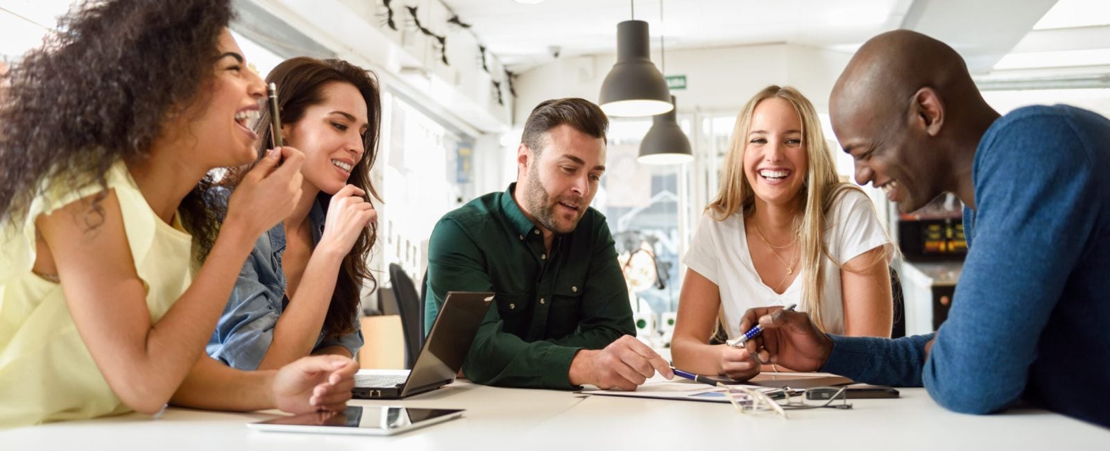 Five young people studying with laptop and tablet computers on white desk. Beautiful girls and guys working toghether wearing casual clothes. Multi-ethnic group smiling.