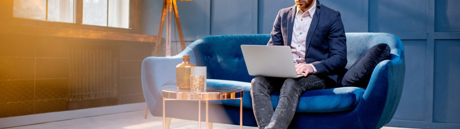 Portrait of an elegant businessman sitting with laptop on the couch at the luxury blue office interior