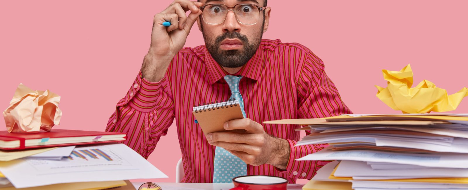 Businessman with scared expression feels frustrated, has finance sheets on desk, holds notepad, drinks coffee, wears formal shirt, isolated over pink background, hears shocking news from office worker