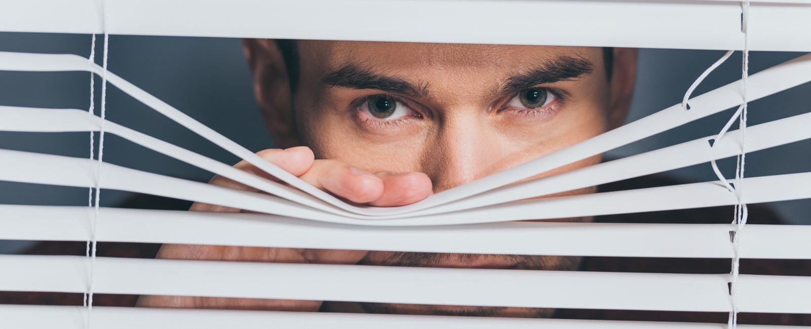 suspicious young man looking at camera through blinds, mistrust concept