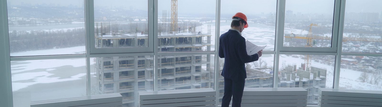 architect holding a design plan or other technical drawing looking on the project . worker in elegant suit and hard hat looking through the windows on the construction site with crane and workers on the building. urban landscape in winter season