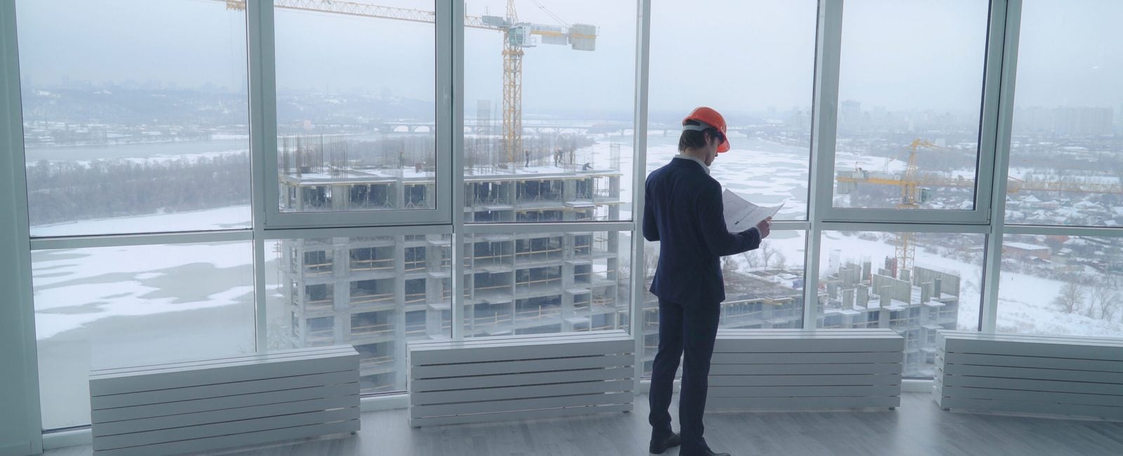architect holding a design plan or other technical drawing looking on the project . worker in elegant suit and hard hat looking through the windows on the construction site with crane and workers on the building. urban landscape in winter season