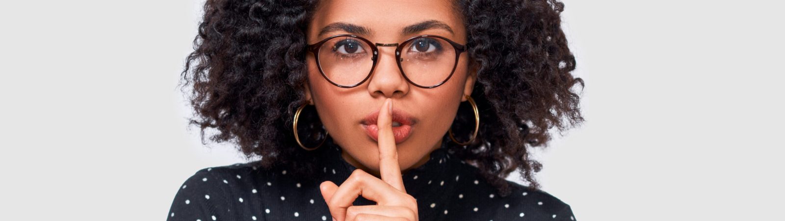 Closeup studio portrait of African American young woman wears black long sleeve shirt, transparent eyewear holding index finger on lips, asking to keep silence. Dark skinned female asks to be quiet.