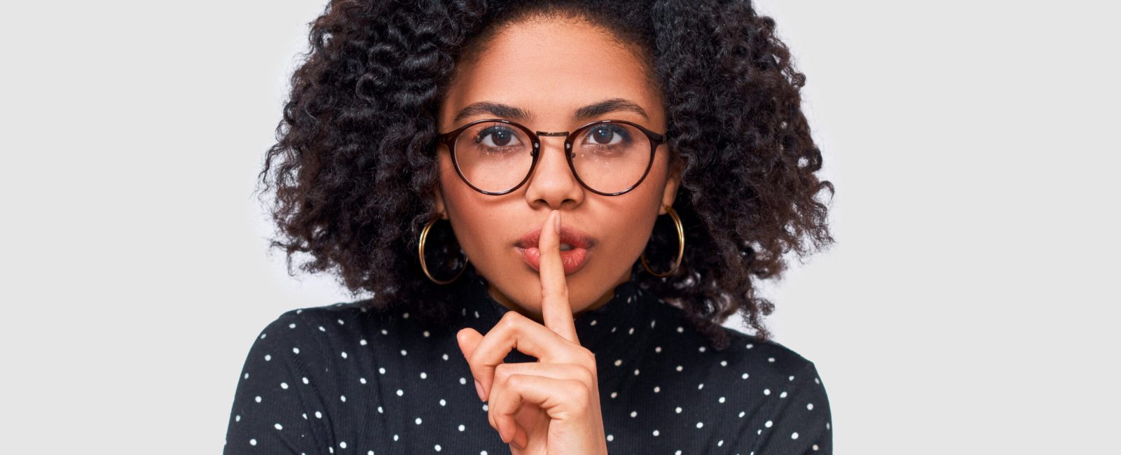 Closeup studio portrait of African American young woman wears black long sleeve shirt, transparent eyewear holding index finger on lips, asking to keep silence. Dark skinned female asks to be quiet.