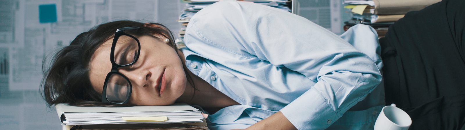 Exhausted business woman sleeping in the office, she is lying down on the desk surrounded by paperwork and empty cups of coffee