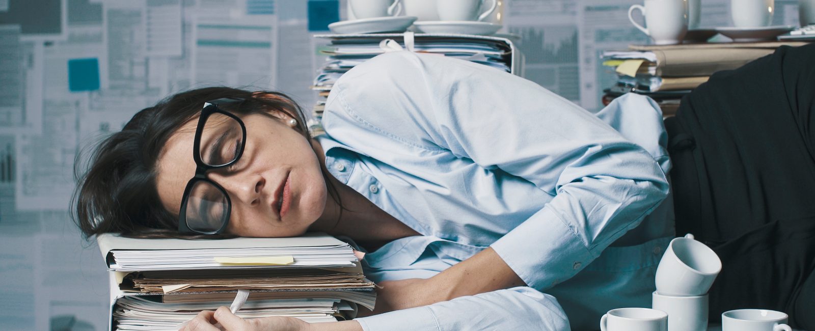 Exhausted business woman sleeping in the office, she is lying down on the desk surrounded by paperwork and empty cups of coffee