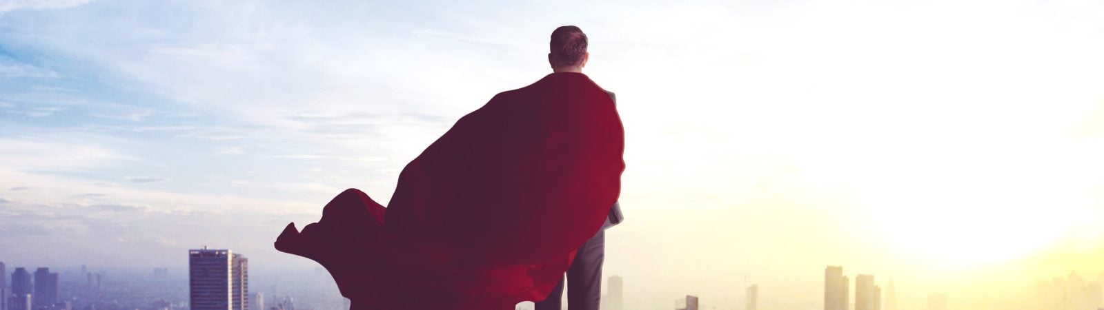 Rear view of young businessman wearing a red cape while standing in the office rooftop and looking at modern city. Shot at morning time