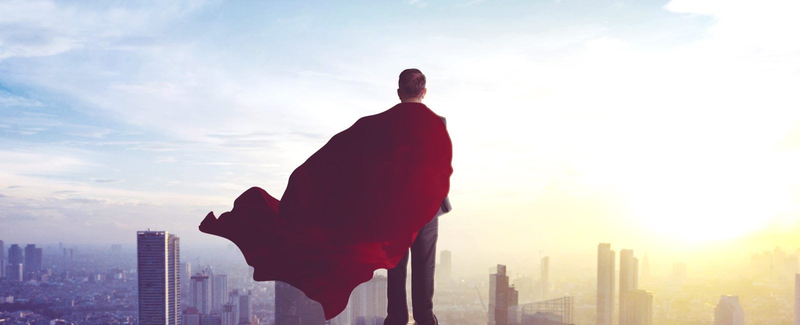 Rear view of young businessman wearing a red cape while standing in the office rooftop and looking at modern city. Shot at morning time