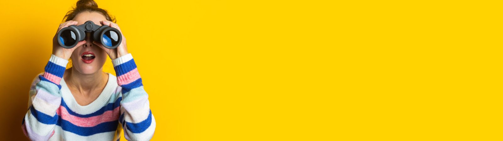 Young woman in sweater looking through binoculars on yellow background.