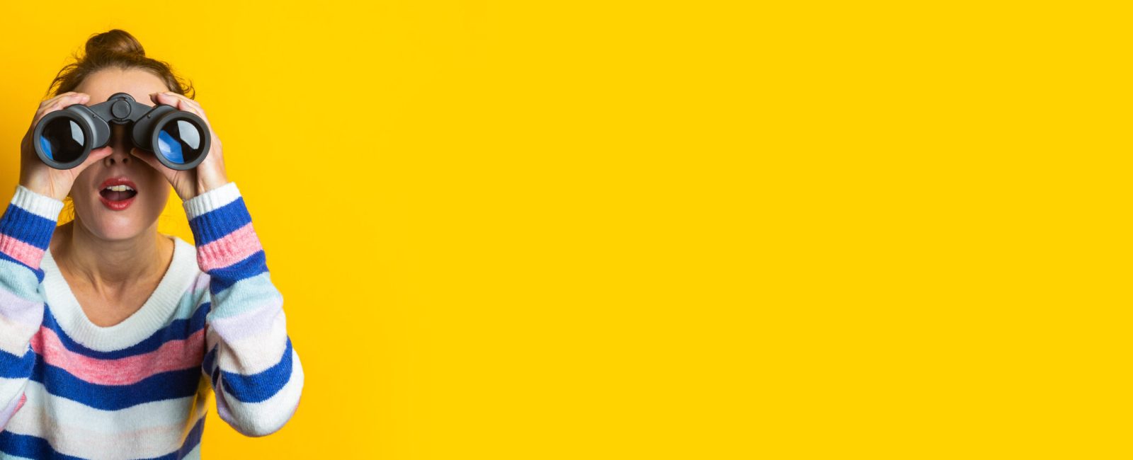 Young woman in sweater looking through binoculars on yellow background.