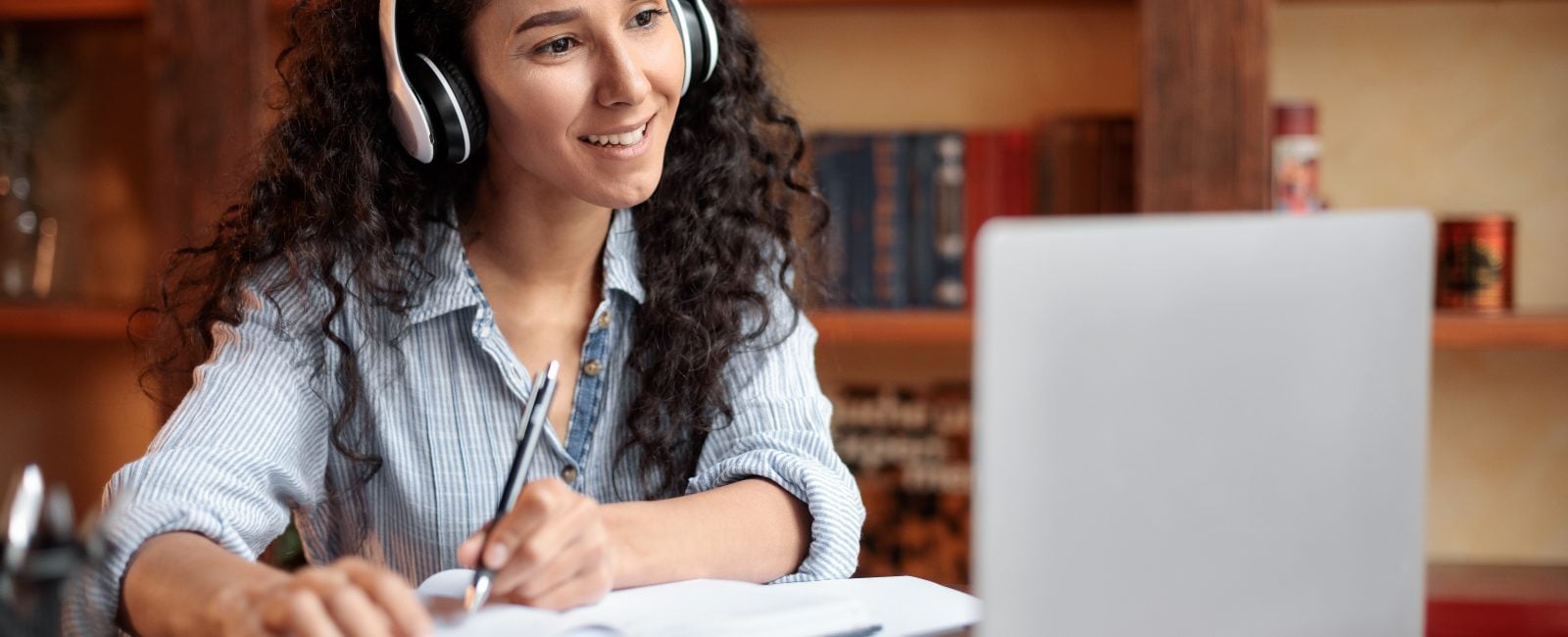 Modern Technology And Elearning. Smiling woman in wireless headset watching webinar online, sitting at desk, using laptop and writing in notebook, taking notes. Education During Quarantine