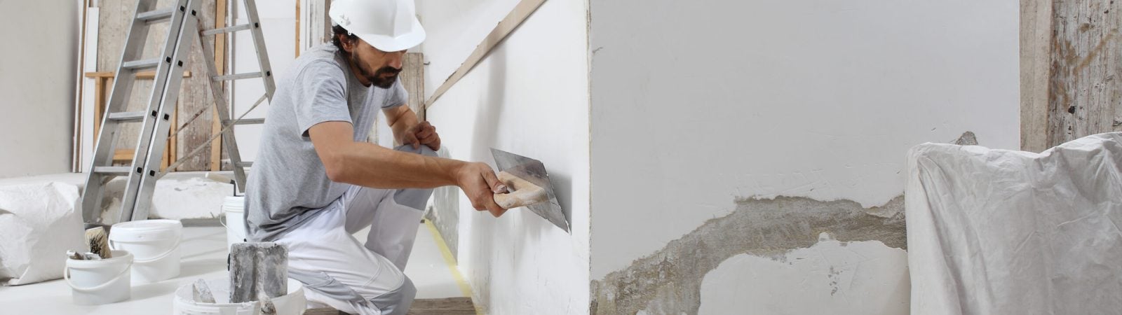 man plasterer construction worker at work, takes plaster from bucket and puts it on trowel to plastering the wall, wears helmet inside the building site of a house
