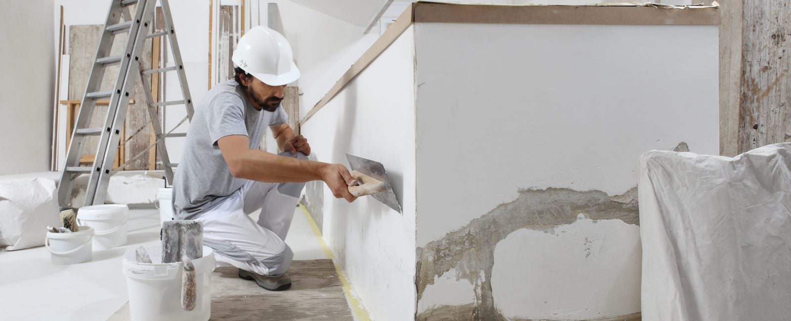 man plasterer construction worker at work, takes plaster from bucket and puts it on trowel to plastering the wall, wears helmet inside the building site of a house