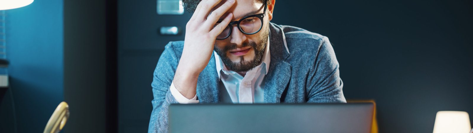 Stressed young businessman sitting at office desk and working with his laptop, he is having a headache
