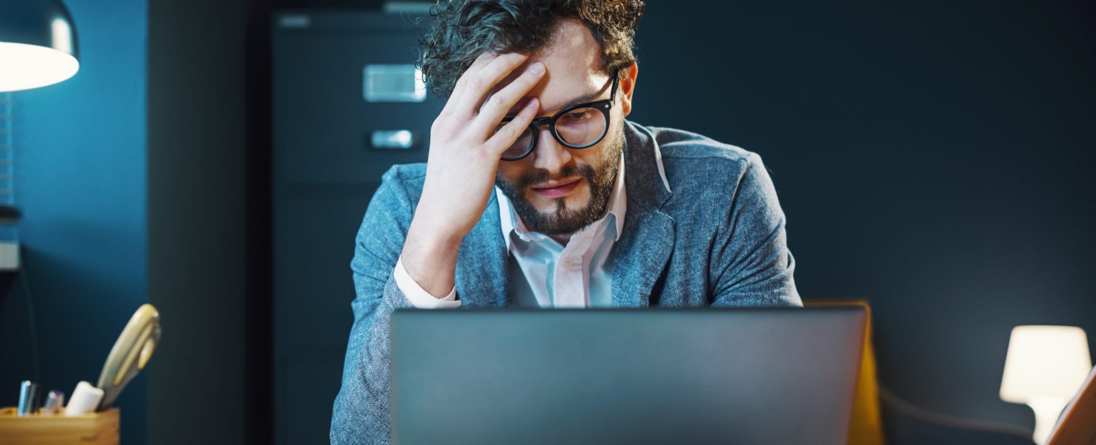 Stressed young businessman sitting at office desk and working with his laptop, he is having a headache