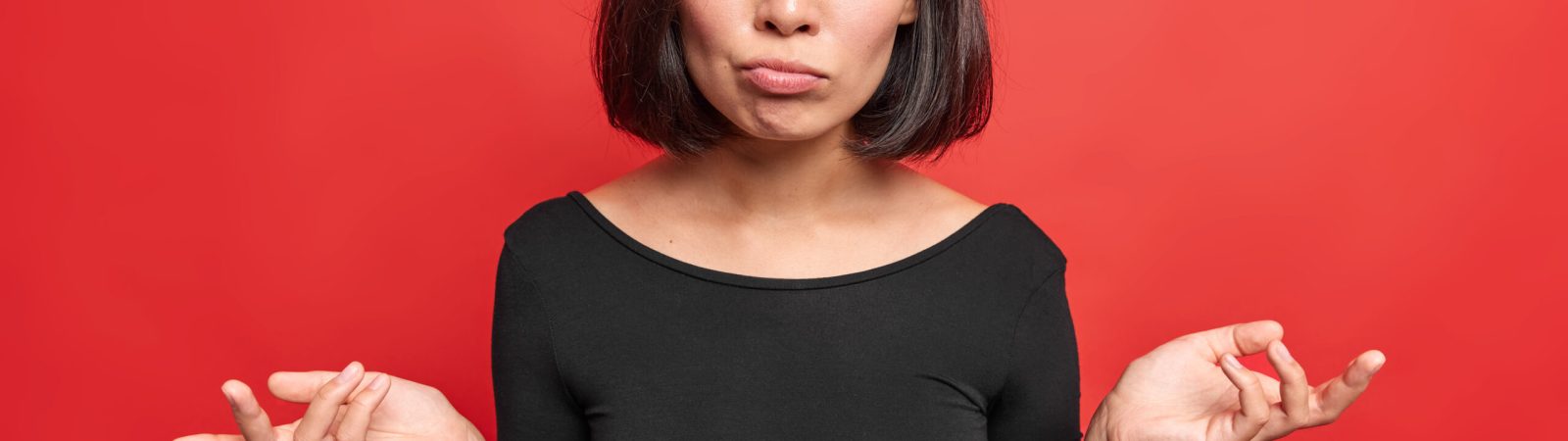 Hesitant confused woman spreads palms looks indignant feels unaware wears black dress raises eyebrows poses against red background shrugs shoulders doesnt know what to do. People uncertainty