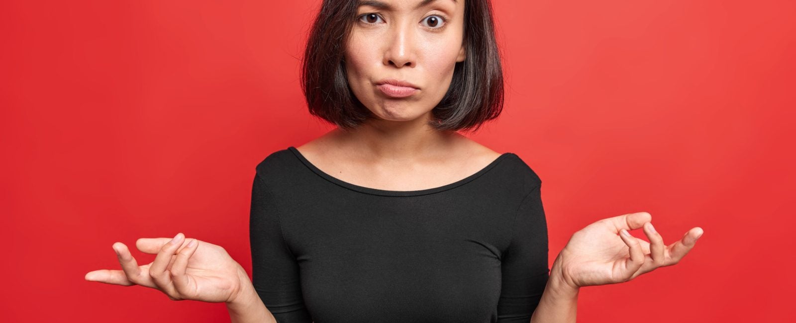 Hesitant confused woman spreads palms looks indignant feels unaware wears black dress raises eyebrows poses against red background shrugs shoulders doesnt know what to do. People uncertainty