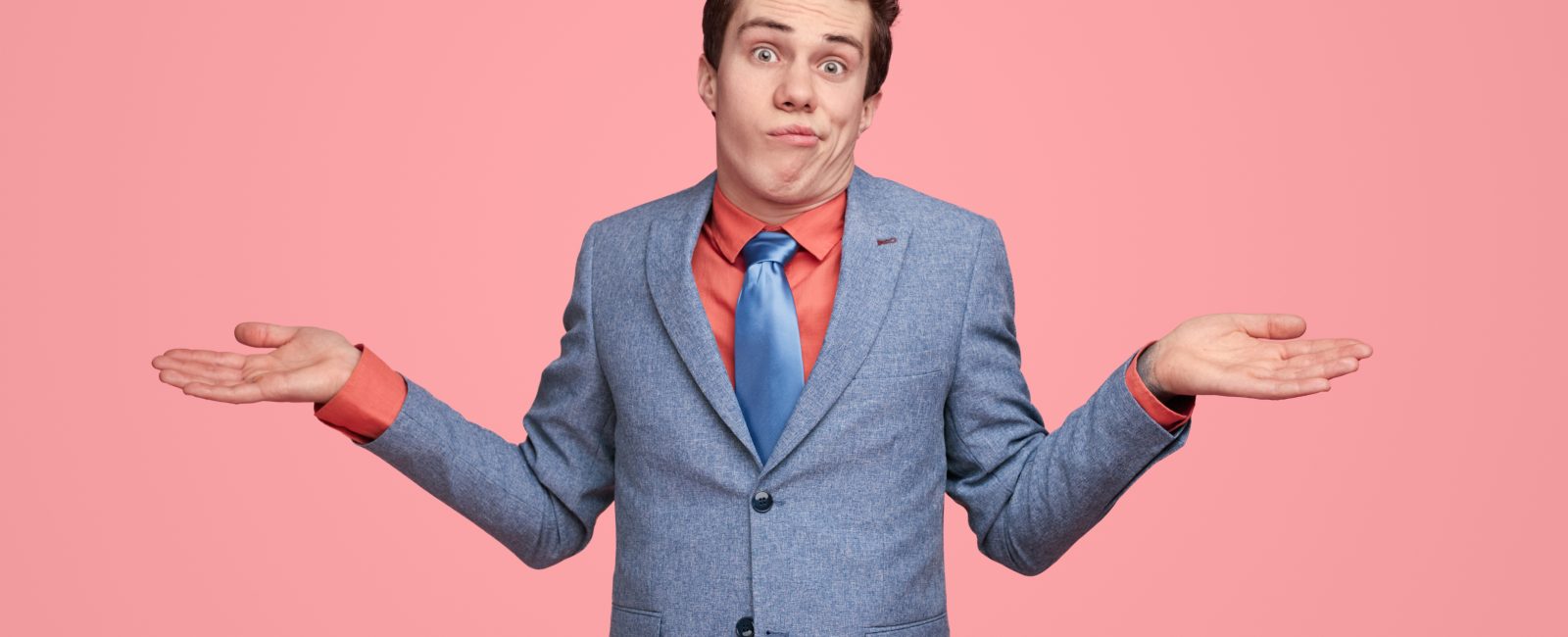 Funny clueless male entrepreneur in suit shrugging shoulders and expressing misunderstanding on pink background in studio while looking at camera