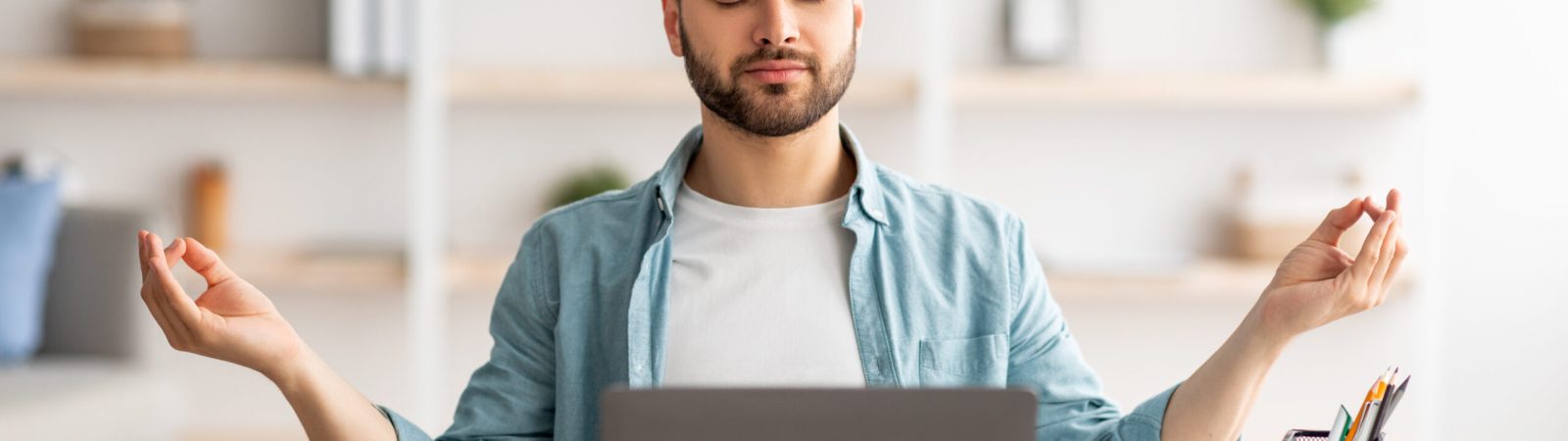 Workplace stress management. Calm Caucasian man meditating with closed eyes in front of laptop pc at home office. Young male freelancer feeling peaceful and balanced, doing yoga at his desk