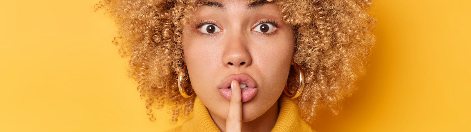 Headshot of surprised curly haired young woman presses index finger over lips makes shush gesture looks mysteriously at camera wears jumper isolated over vivid yellow background tells secret
