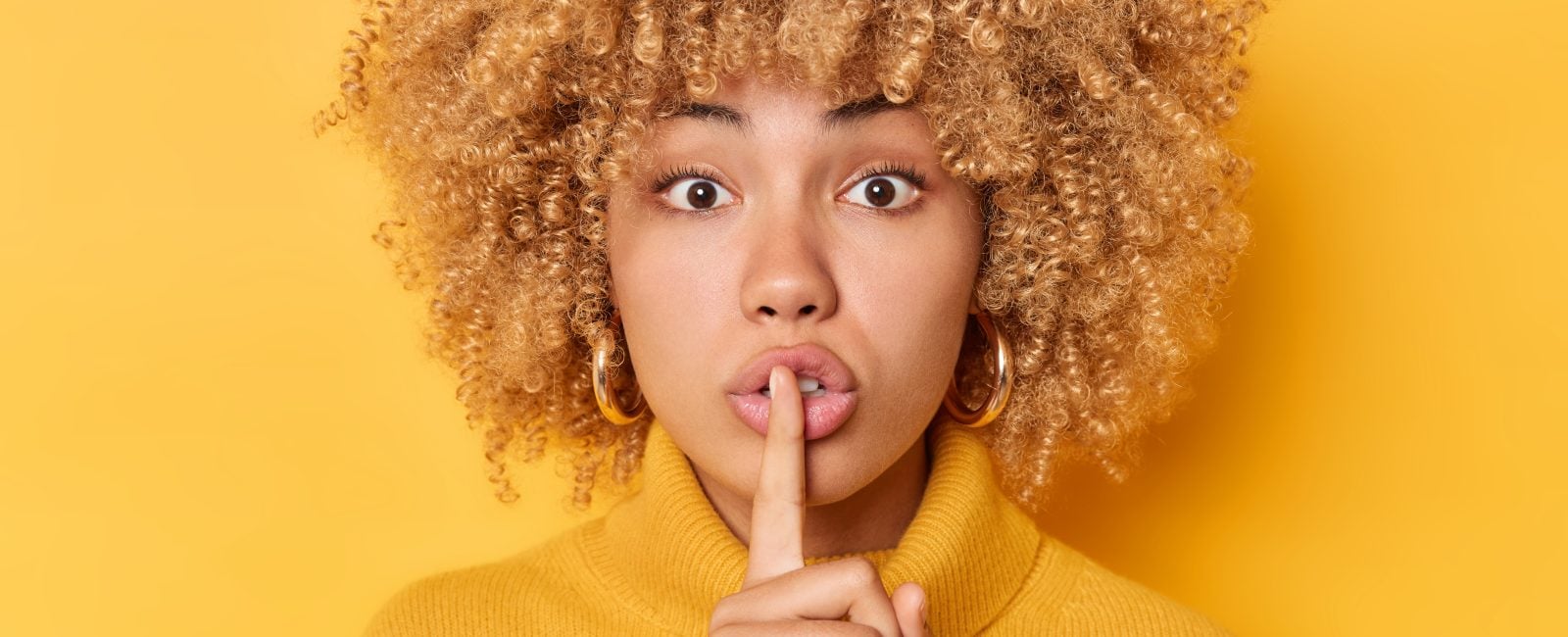 Headshot of surprised curly haired young woman presses index finger over lips makes shush gesture looks mysteriously at camera wears jumper isolated over vivid yellow background tells secret