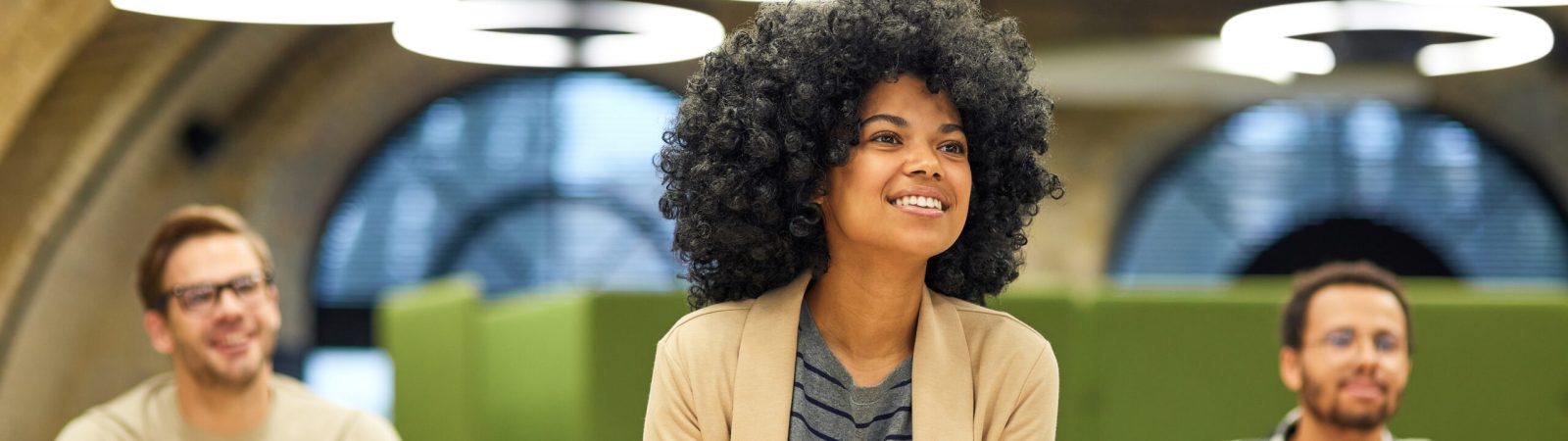 Happy mixed race woman studying with colleagues while sitting in coworking space, listening to coach during corporate training or seminar. Business education concept