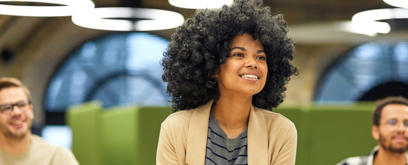 Happy mixed race woman studying with colleagues while sitting in coworking space, listening to coach during corporate training or seminar. Business education concept