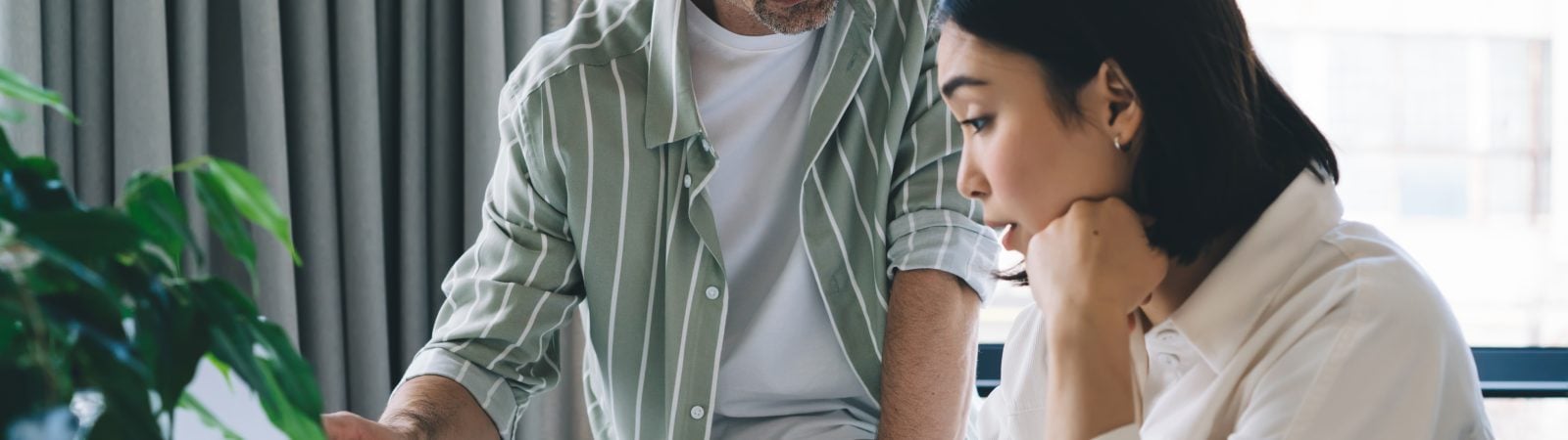 Side view of young Asian female employee sitting at table while taking notes and paying attention to manager explain office project using laptop