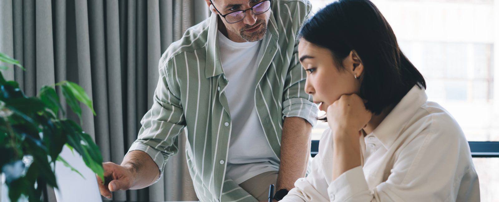 Side view of young Asian female employee sitting at table while taking notes and paying attention to manager explain office project using laptop